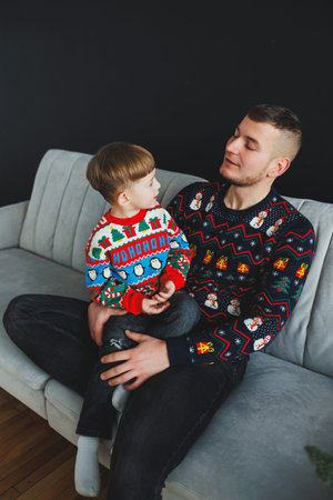 A heartwarming moment captured with a father and son wearing colorful holiday sweaters, sharing smiles on a cozy couch. Perfect for celebrating family traditions.の写真素材