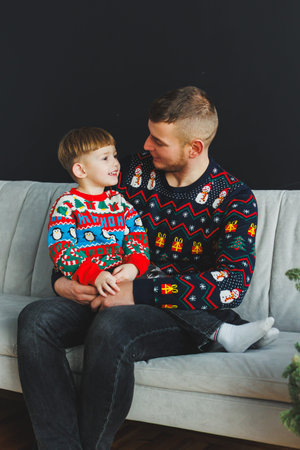 A joyful father and son share a heartwarming moment on the couch, both in colorful holiday sweaters, celebrating the spirit of togetherness this festive seasonの写真素材