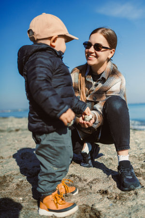 A woman and a child are on the beach. The woman is smiling as she kneels down and holds the child's hands. They are enjoying the sunny weather by the water.の写真素材