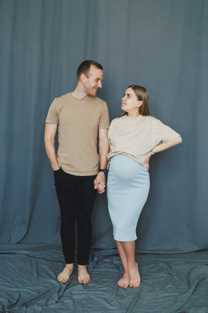 A man and a woman are holding hands in a studio. The woman shows her baby bump, and both are smiling at each other. They stand on a blue fabric floor.の写真素材