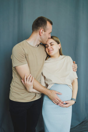 A man kisses a woman on the forehead as they pose together for a maternity session. The woman rests her hands on her belly while smiling. They stand in a studio with a blue background.の写真素材