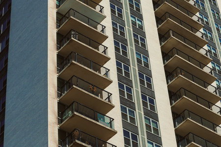 A tall apartment building stands against a clear blue sky. The structure features multiple balconies and many windows, showing signs of urban life. People may be seen enjoying their spaces.の写真素材
