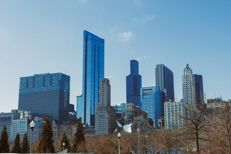 People walk and enjoy the sunny day in a park while looking at the tall buildings in Chicago. The skyline shows different shapes and heights of buildings against the blue sky.の写真素材