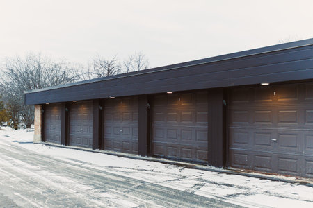 A row of closed garage doors is seen in a snowy area. Ice is on the ground, and bare trees are in the background. The scene shows the quiet of winter in a suburban setting.の写真素材