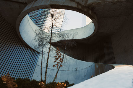 A spiral concrete path leads upwards, framed by tall buildings and trees. The sky is clear, and sunlight illuminates the scene. The structure shows modern design elements and urban nature.の写真素材