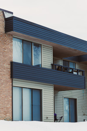 Two-story building with balconies and large windows shows signs of winter with snow covering the ground and some trees without leaves in a neighborhood during a cloudy day.の写真素材