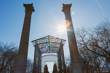 Two tall columns stand beside an arch structure in a park. Trees without leaves surround the area. Bright sun shines in a clear blue sky at noon.の写真素材