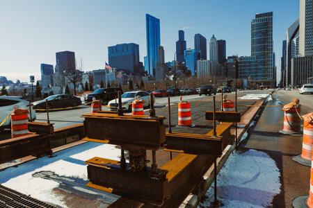 Construction equipment and barriers are set up along a city road in Chicago. Cars pass by while workers maintain the area under a clear sky. The buildings stand tall in the background.の写真素材