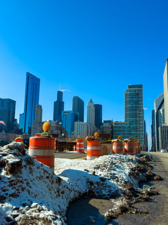 Construction barriers are set up along the street with snow piles in Chicago. Tall buildings rise in the background under a bright blue sky during early spring.の写真素材