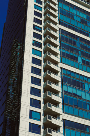 A tall modern building stands in the city with large glass windows and multiple balconies. Sunlight...