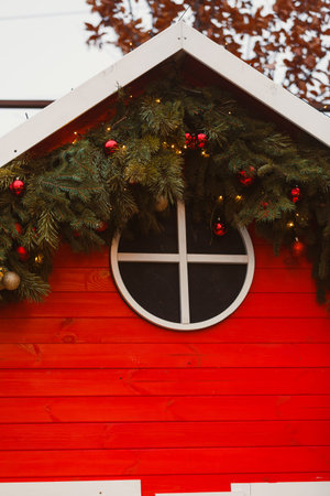 A red wooden house features a round window and is decorated with green branches and shiny ornaments. The scene shows parts of the roof under bright sunlight during winter time.の写真素材