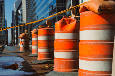 Workers set up construction barriers on a city street lined with tall buildings. Orange barrels and caution tape keep pedestrians away from the work area. It is daytime, and the street is busy.の写真素材