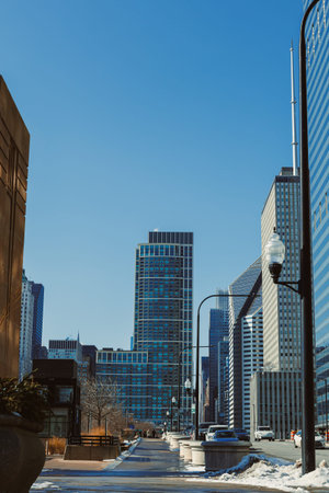 People walk along the street surrounded by tall buildings in a city. The sky is blue with no clouds, and trees and benches are seen nearby.の写真素材