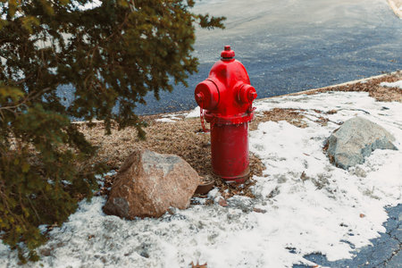 A red fire hydrant sits beside some rocks and snow in a winter scene. It is located near a road and...