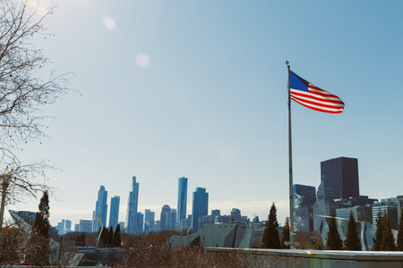 A clear day shows a view of tall buildings in the city. An American flag flutters in the foreground against the skyline. Trees and rooftops are visible below the buildings.の写真素材