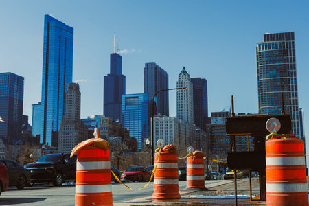 Barriers block the street in downtown Chicago. Skyscrapers rise above the city. Cars are parked along the road. The sky is clear and blue. Workers are likely nearby.の写真素材