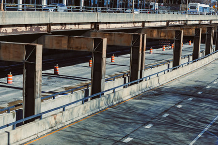 Construction activities occur on a highway underpass with orange cones set up. Trucks and vehicles...