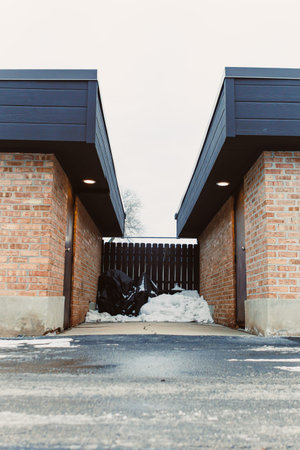 In a residential area, snow is piled against a wooden fence surrounded by two brick walls. The ground shows signs of winter's chill with icy surfaces and a gray sky above.の写真素材