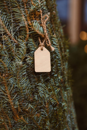 A wooden tag hangs from a Christmas tree branch. The tree has a rich green color. This scene takes place in a festive environment with lights and decorations for the holiday season.の写真素材