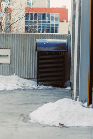 A trash can stands against a wall in a winter scene. Snow covers the ground while buildings rise in the background. This setting shows a typical urban environment during cold weather.の写真素材