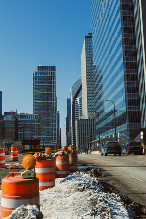 Workers set up construction barriers during the day on a street filled with tall buildings. Vehicles...