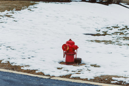A red fire hydrant is visible on a patch of snow-covered grass next to a black paved road in a residential neighborhood during winter.の写真素材