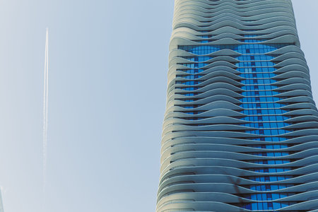 A tall building features a unique wavy structure and large glass windows. A plane leaves a trail in the sky above the urban setting during bright daylight.の写真素材