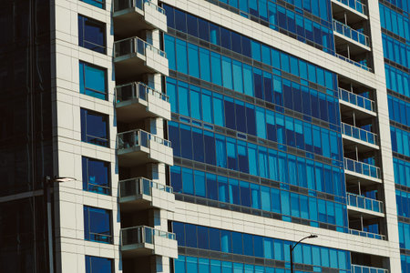 A high-rise building stands tall with many glass windows. The structure features balconies that overlook the city streets. The sunlight reflects off the blue glass, creating a bright scene.の写真素材
