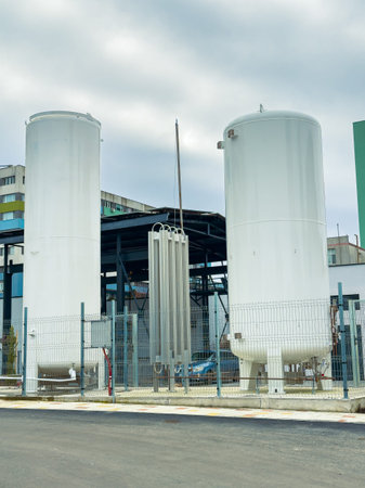 Storage tanks are placed beside a building in an industrial site. The tanks are tall and white, and there are steel pipes and a fence nearby. Cloudy skies are above.の写真素材