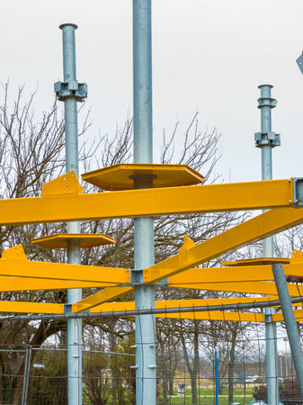 Workers are constructing a playground structure with yellow components in a park. Metal poles rise...