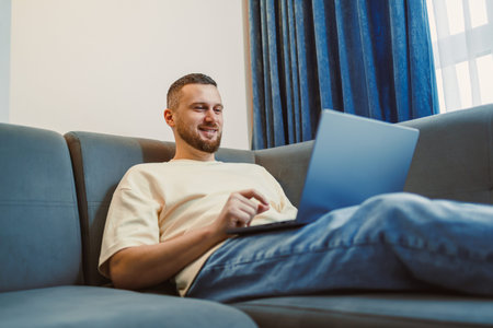 A man sits on a sofa and uses a laptop in a living room. Sunlight comes through the window. He appears engaged and content with his work.の写真素材