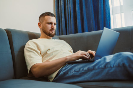 A man is seated on a couch using a laptop in a stylish living room. He shows focus as he types....