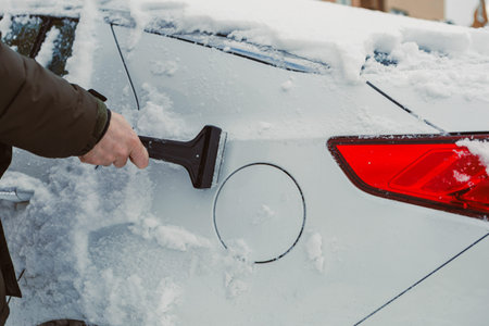 A person is removing snow from the back of a car using a scraper. The scene shows a winter day with...