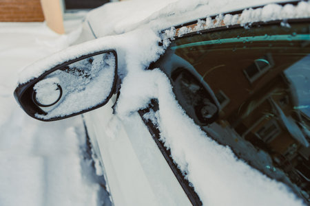 Snow is piled on a parked car in a neighborhood. The car's side mirror is covered in white frost....