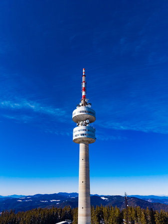 A television tower rises above the mountains under a clear blue sky. Trees surround the base, and snow can be seen on the ground. The scene shows the tower's design and location in nature.の写真素材