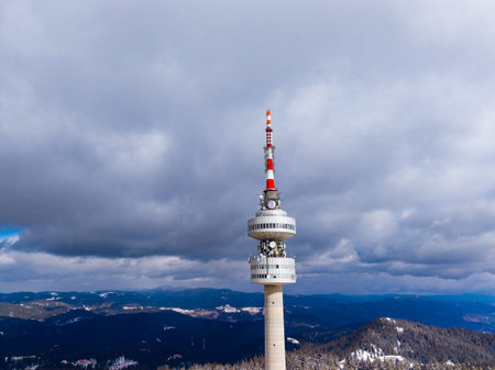 The scene shows a tall communication tower surrounded by clouds and mountains. The tower is in a...