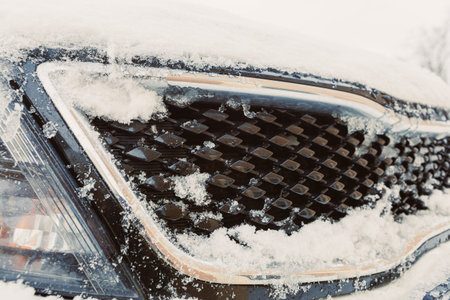 The front of a car shows a grille covered in snow. Snowflakes fall around the vehicle. The scene shows the effects of winter weather on a car parked outside.の写真素材