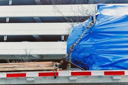 A truck is parked at a construction site with a blue tarp covering wooden materials. It is near a parking structure on a clear day with no people visible.の写真素材