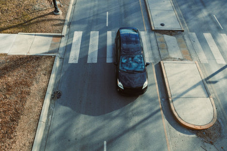 A black car moves across a crosswalk on a busy street in an urban area. Trees and buildings create...
