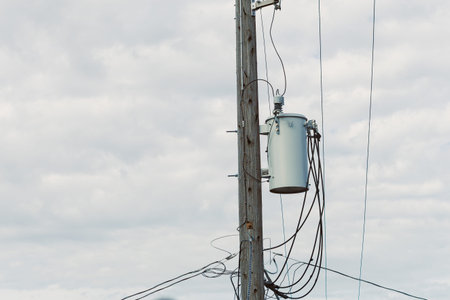 A wooden utility pole stands tall with a transformer attached to it. Wires hang down from the transformer. Clouds cover the sky overhead.の写真素材