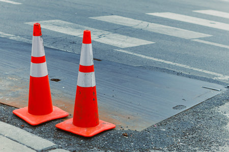Traffic cones stand on the side of a city street near a marked crosswalk. The ground shows signs of...
