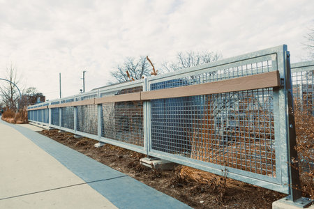 Individuals stroll along a path next to a tall metal fence in a city setting under a cloudy sky....