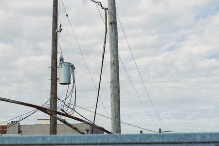 Power lines and wooden poles rise into the sky against a backdrop of clouds. Utility equipment is...
