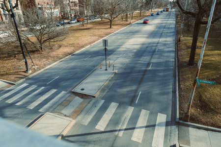 A street in an urban area shows empty lanes with a few parked cars. The sky is clear, and the scene...