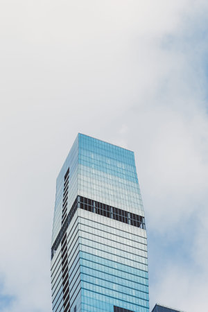 A tall building made of glass rises against the sky with clouds around it. Light reflects off the...