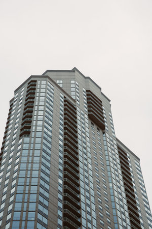 View from below of a tall building with many windows set in an urban space under a cloudy sky.