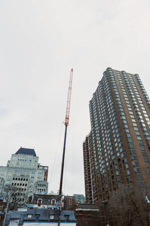 A construction crane reaches high into the sky between tall buildings in a city. The ground is busy with activity as workers manage their tasks below.の写真素材
