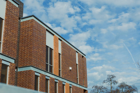 A brick building stands with multiple windows. The sky is blue and has clouds. The setting is during...