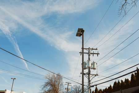 Power lines stretch across the sky in a suburban area. The clear blue sky has clouds and sunlight....
