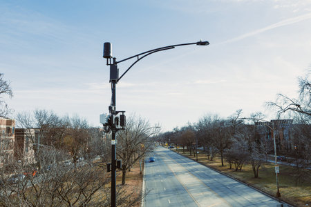 A traffic camera is fixed to a streetlight on a quiet road. The road is bordered by bare trees and...